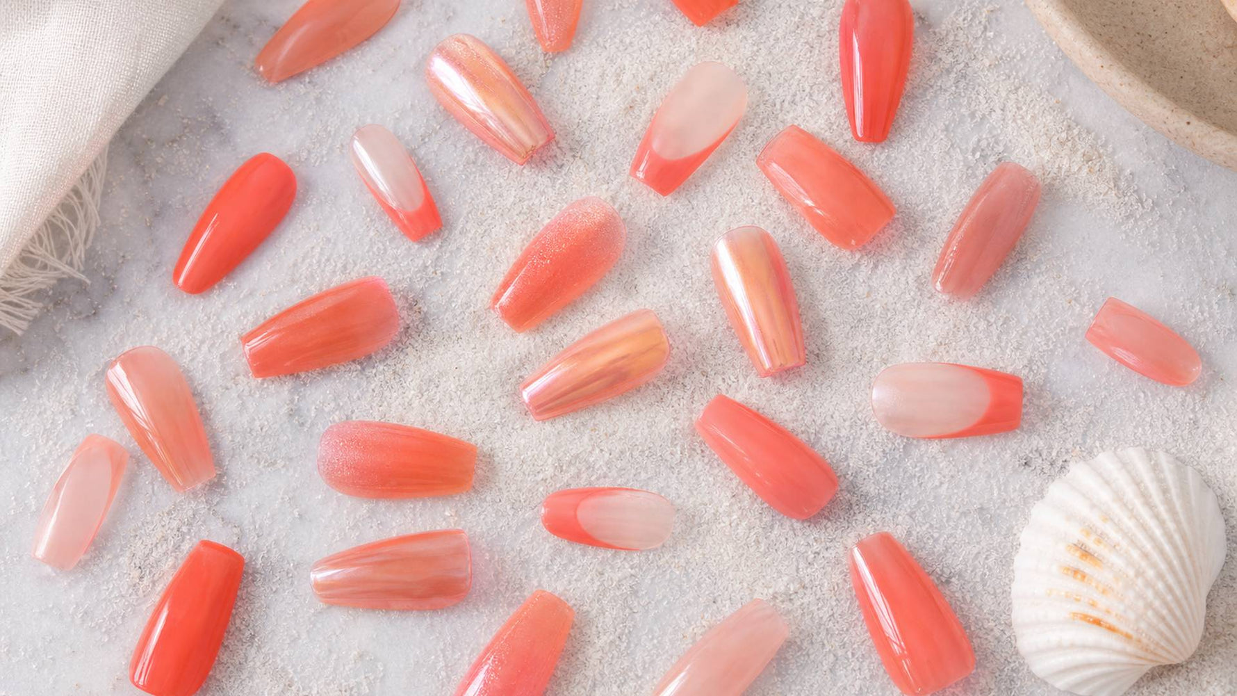 coral press-on nails flat lay showing bright coral, soft coral, French tip, and chrome designs on almond and coffin shapes against a white sandy summer background